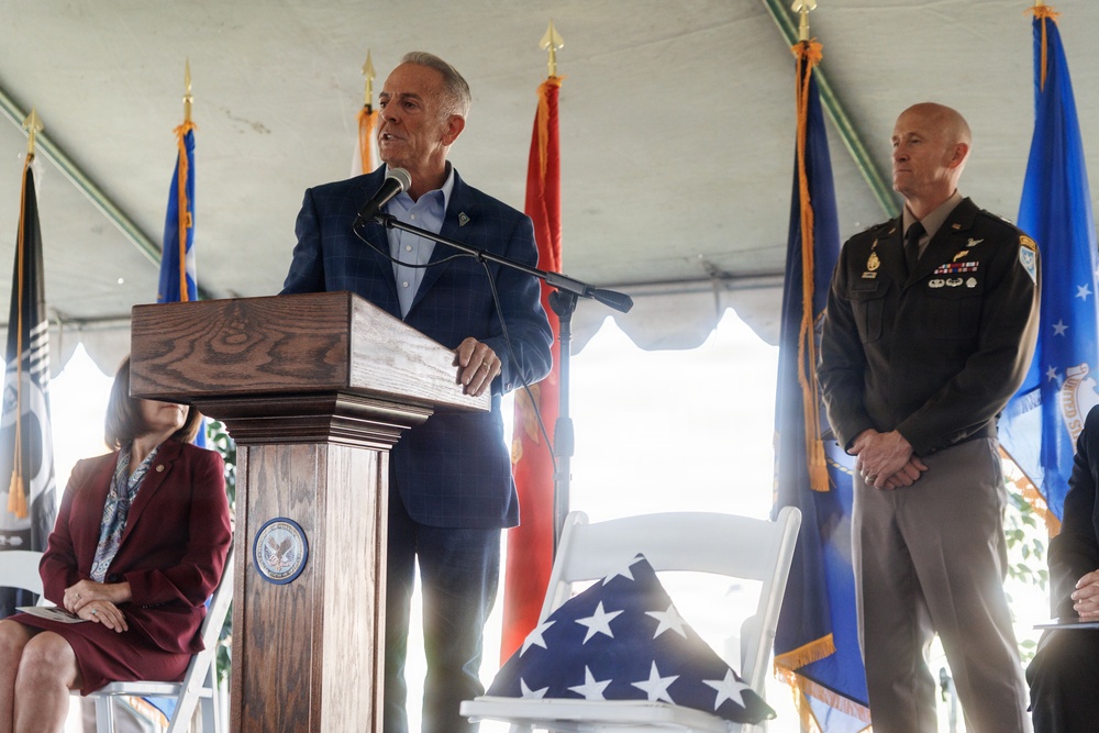 A formal military or veterans ceremony is taking place under a white tent. A civilian man in a blue blazer is delivering remarks at a wooden podium bearing an official seal, flanked by a military officer in Army dress uniform and a seated woman in a red blazer. Multiple military branch flags and a folded American flag on a chair are prominently displayed.