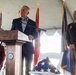 A formal military or veterans ceremony is taking place under a white tent. A civilian man in a blue blazer is delivering remarks at a wooden podium bearing an official seal, flanked by a military officer in Army dress uniform and a seated woman in a red blazer. Multiple military branch flags and a folded American flag on a chair are prominently displayed.