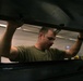 A young man wearing an olive/army green t-shirt is working on what appears to be a military vehicle, likely a HMMWV (Humvee), in an indoor garage or motor pool facility. He is reaching upward with both hands to manipulate a panel or hatch mechanism above him. The image is shot from a low angle through an opening in the vehicle, creating a dramatic framing effect.