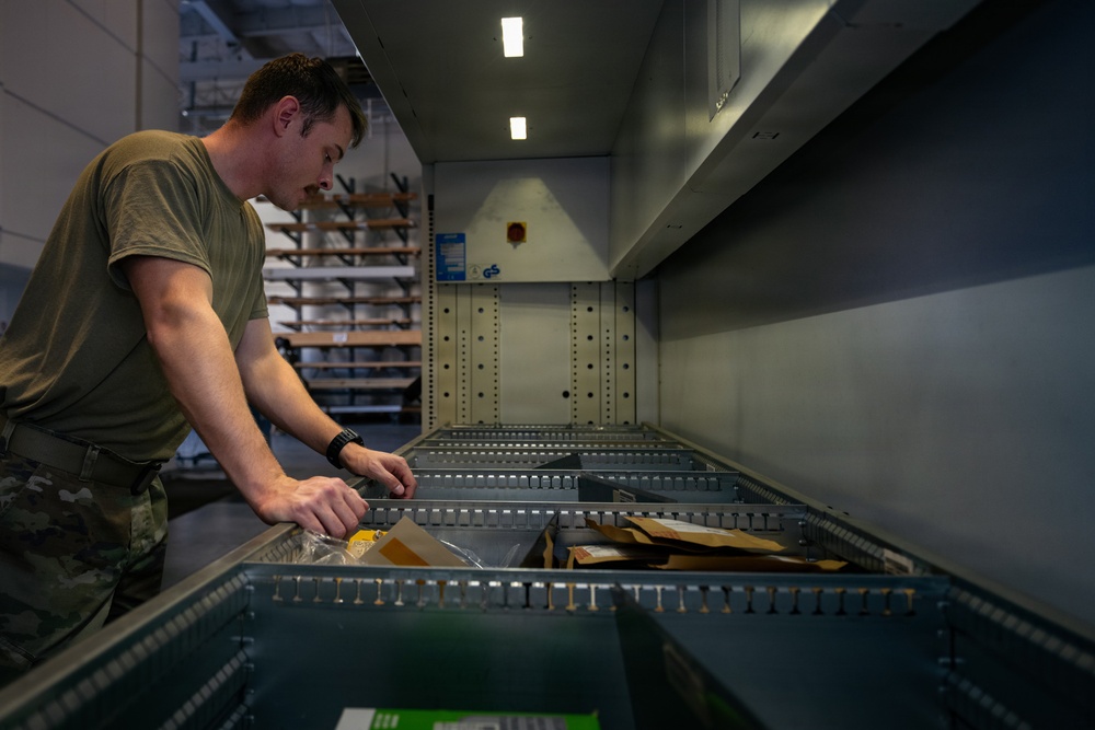 A military service member in olive drab/camouflage uniform is working in what appears to be a military supply or mail room facility. He is leaning over a metal shelving or sorting unit, handling packages and envelopes. The setting suggests a logistics, postal, or supply operations environment.