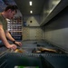 A military service member in olive drab/camouflage uniform is working in what appears to be a military supply or mail room facility. He is leaning over a metal shelving or sorting unit, handling packages and envelopes. The setting suggests a logistics, postal, or supply operations environment.