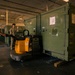 Two military personnel in camouflage uniforms are working inside a warehouse or storage facility, maneuvering large olive-green military containers using a yellow electric pallet jack. One soldier operates the pallet jack while the other appears to be directing or guiding the movement. The containers are labeled with alphanumeric identifiers and have documentation attached to them.