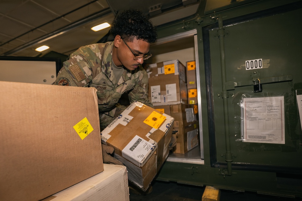 A U.S. military service member in Army Combat Uniform (ACU) with Operational Camouflage Pattern (OCP) is loading or unloading cardboard boxes and packages into a green military storage container or aircraft cargo hold. The packages include various shipping labels, yellow 'Attention' warning stickers, and appear to be military spare parts or supplies. The setting appears to be inside a cargo aircraft or military logistics facility.
