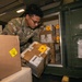 A U.S. military service member in Army Combat Uniform (ACU) with Operational Camouflage Pattern (OCP) is loading or unloading cardboard boxes and packages into a green military storage container or aircraft cargo hold. The packages include various shipping labels, yellow 'Attention' warning stickers, and appear to be military spare parts or supplies. The setting appears to be inside a cargo aircraft or military logistics facility.