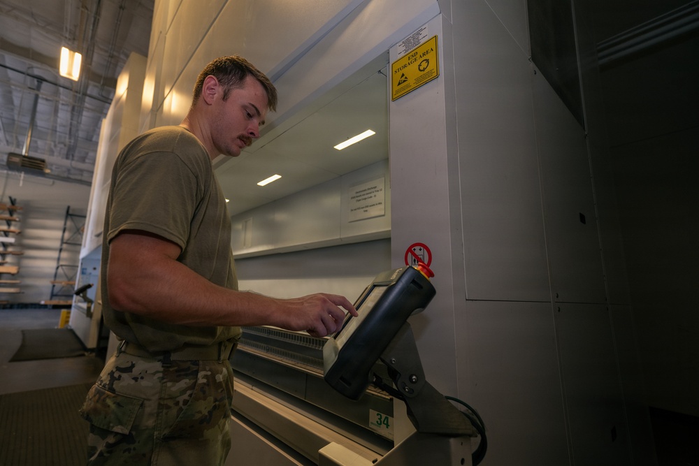 A military service member in camouflage uniform and olive drab t-shirt operates what appears to be a large-format industrial machine or printer in a facility. The person is interacting with a control panel or touchscreen interface on the equipment. The setting appears to be a military maintenance or technical facility.