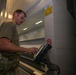 A military service member in camouflage uniform and olive drab t-shirt operates what appears to be a large-format industrial machine or printer in a facility. The person is interacting with a control panel or touchscreen interface on the equipment. The setting appears to be a military maintenance or technical facility.