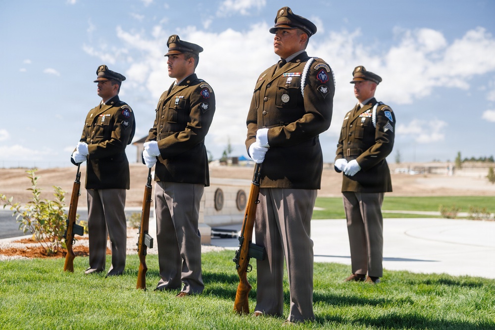 Four U.S. Army soldiers in dress green uniforms stand at attention on a grass lawn, appearing to participate in a military honor guard ceremony. Three of the soldiers are holding rifles, while one stands with hands clasped in front. The setting appears to be a military cemetery or memorial grounds based on the manicured landscape and ceremonial context.