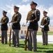 Four U.S. Army soldiers in dress green uniforms stand at attention on a grass lawn, appearing to participate in a military honor guard ceremony. Three of the soldiers are holding rifles, while one stands with hands clasped in front. The setting appears to be a military cemetery or memorial grounds based on the manicured landscape and ceremonial context.