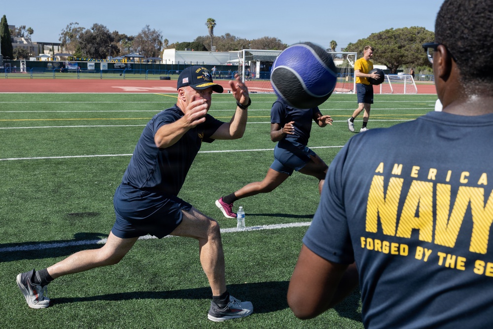 A group of U.S. Navy personnel are engaged in an outdoor physical training session on a green turf athletic field. The foreground shows individuals passing or catching a large medicine ball, with one person lunging forward to receive it. The setting appears to be a military fitness exercise at a sports complex with a running track and soccer goal visible in the background.