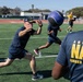 A group of U.S. Navy personnel are engaged in an outdoor physical training session on a green turf athletic field. The foreground shows individuals passing or catching a large medicine ball, with one person lunging forward to receive it. The setting appears to be a military fitness exercise at a sports complex with a running track and soccer goal visible in the background.