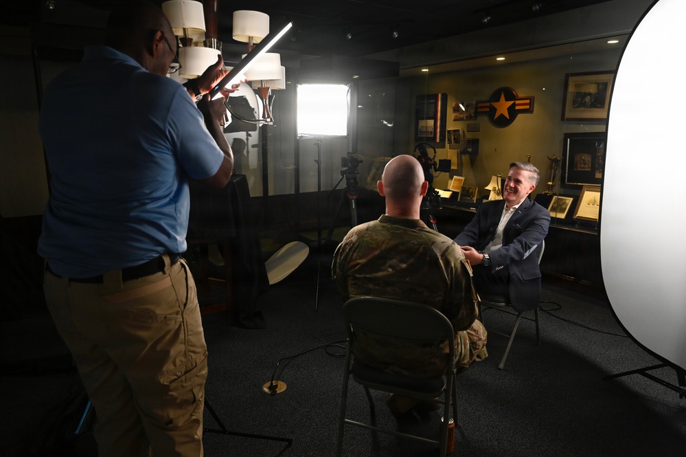 A professional video or photo interview is being conducted in what appears to be a military office or briefing room. A man in a suit is being interviewed by a person in military camouflage uniform, while a civilian crew member holds a lighting panel to illuminate the scene. The background features military aviation insignia, framed photographs, and decorative items consistent with a senior military official's office.