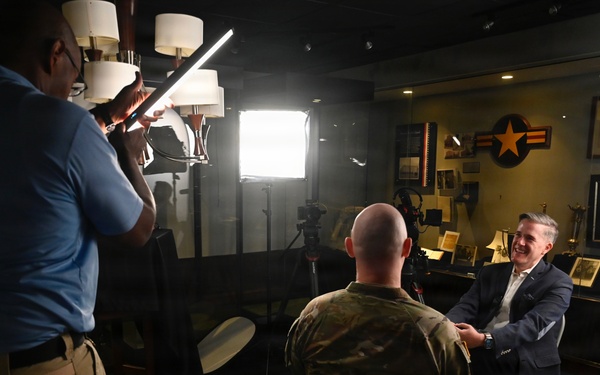 A professional video or photo interview is being conducted in what appears to be a military office or briefing room. A man in a suit is being interviewed by a person in military camouflage uniform, while a civilian crew member holds a lighting panel to illuminate the scene. The background features military aviation insignia, framed photographs, and decorative items consistent with a senior military official's office.