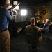 A professional video or photo interview is being conducted in what appears to be a military office or briefing room. A man in a suit is being interviewed by a person in military camouflage uniform, while a civilian crew member holds a lighting panel to illuminate the scene. The background features military aviation insignia, framed photographs, and decorative items consistent with a senior military official's office.
