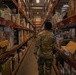 A person wearing military camouflage (OCP/MultiCam pattern) uniform is walking down an aisle in a large military supply warehouse or logistics facility. The warehouse contains tall metal shelving units filled with boxes, packages, and various supply items. The image is taken from behind the individual, showing the depth of the storage aisle receding into the background.