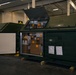 The image shows a large dark green military or industrial storage container, labeled '3A' and '3B', opened to reveal multiple cardboard boxes with orange labels inside. The container is located in what appears to be a warehouse or logistics facility with fluorescent lighting. Documents are affixed to the exterior panel of the container.