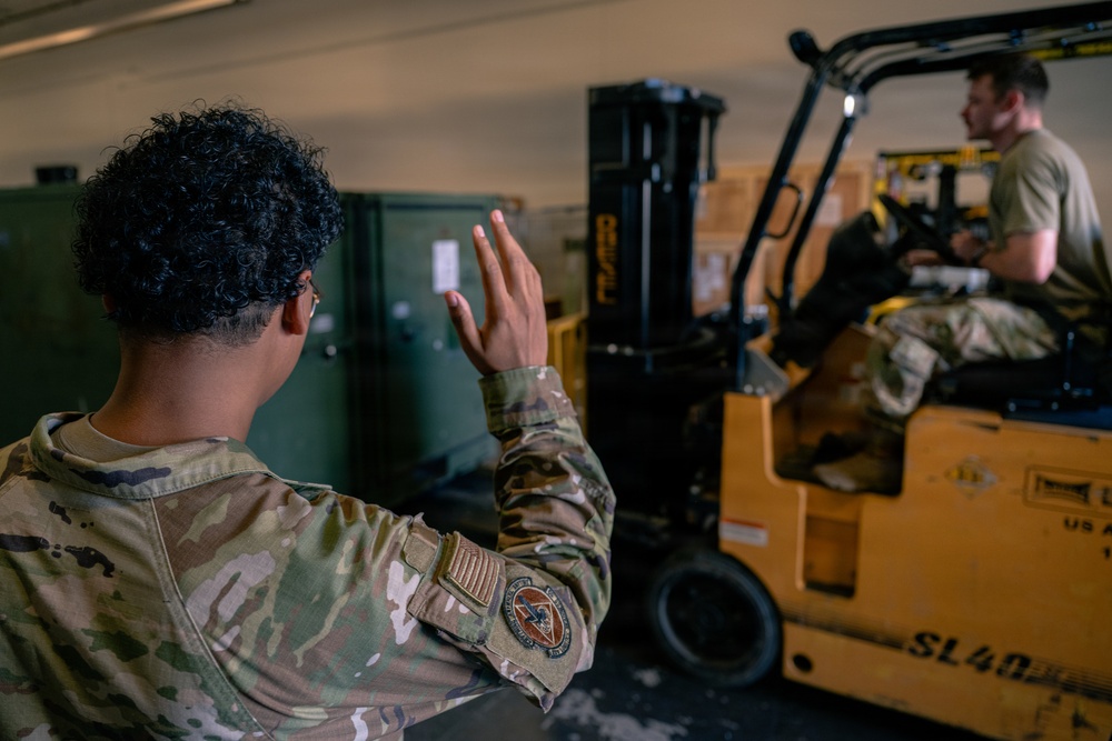 Two military personnel in OCP (Operational Camouflage Pattern) uniforms are working in what appears to be a warehouse or cargo bay. One soldier is directing with a raised hand signal while another operates a yellow forklift marked 'SL40' with 'US' markings, indicating US military property. Cardboard boxes and green military containers are visible in the background.