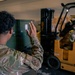 Two military personnel in OCP (Operational Camouflage Pattern) uniforms are working in what appears to be a warehouse or cargo bay. One soldier is directing with a raised hand signal while another operates a yellow forklift marked 'SL40' with 'US' markings, indicating US military property. Cardboard boxes and green military containers are visible in the background.