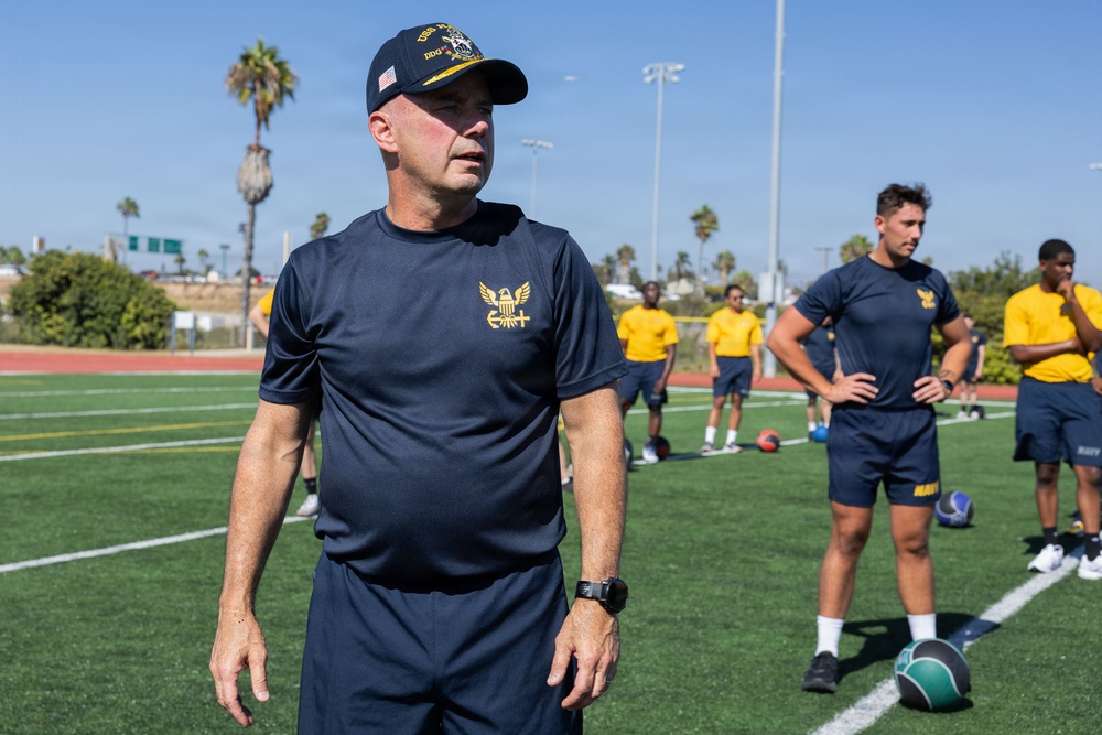 A group of individuals wearing U.S. Navy physical training (PT) uniforms are gathered on an athletic field with artificial turf. The man in the foreground wears a dark navy PT shirt with a gold eagle insignia and a Navy ball cap marked 'DDG' with a U.S. flag patch. The setting appears to be an outdoor sports facility, likely during a physical fitness or training exercise.