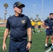 A group of individuals wearing U.S. Navy physical training (PT) uniforms are gathered on an athletic field with artificial turf. The man in the foreground wears a dark navy PT shirt with a gold eagle insignia and a Navy ball cap marked 'DDG' with a U.S. flag patch. The setting appears to be an outdoor sports facility, likely during a physical fitness or training exercise.