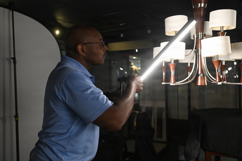 A man wearing glasses and a blue polo shirt is holding a bright LED light tube or wand, directing it toward a decorative chandelier in what appears to be a photography studio or showroom. The setting includes a white reflector panel on the left and a multi-arm chandelier with white shades and wooden accents on the right. The scene suggests a product photography or lighting setup session.