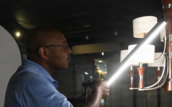 A man wearing glasses and a blue polo shirt is holding a bright LED light tube or wand, directing it toward a decorative chandelier in what appears to be a photography studio or showroom. The setting includes a white reflector panel on the left and a multi-arm chandelier with white shades and wooden accents on the right. The scene suggests a product photography or lighting setup session.