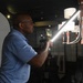 A man wearing glasses and a blue polo shirt is holding a bright LED light tube or wand, directing it toward a decorative chandelier in what appears to be a photography studio or showroom. The setting includes a white reflector panel on the left and a multi-arm chandelier with white shades and wooden accents on the right. The scene suggests a product photography or lighting setup session.