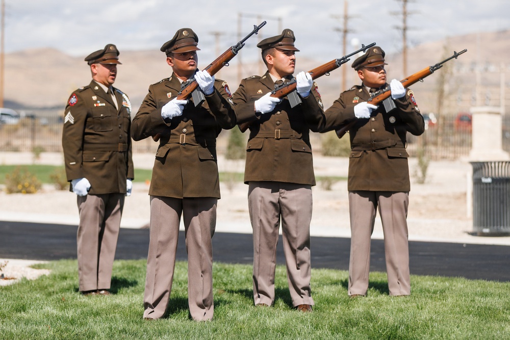 A military honor guard ceremony is taking place outdoors, with three rifle bearers in olive drab dress uniforms raising their rifles in a firing salute position. A fourth uniformed soldier stands at attention in the background, appearing to be the commanding NCO. The setting appears to be a military installation or cemetery in an arid, desert-like environment.