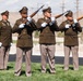 A military honor guard ceremony is taking place outdoors, with three rifle bearers in olive drab dress uniforms raising their rifles in a firing salute position. A fourth uniformed soldier stands at attention in the background, appearing to be the commanding NCO. The setting appears to be a military installation or cemetery in an arid, desert-like environment.
