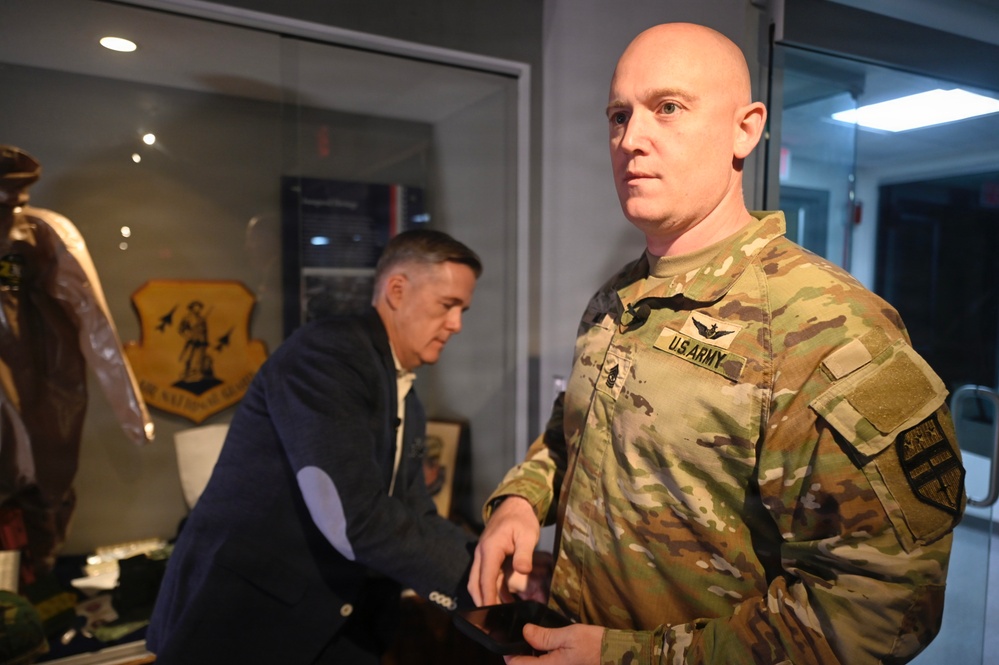 A U.S. Army soldier in OCP (Operational Camouflage Pattern) uniform stands in the foreground inside what appears to be a military heritage or display room. A second individual in civilian attire is visible in the background, leaning over a display table. Military memorabilia, including a leather flight jacket and a unit shield/crest, are visible in the background.