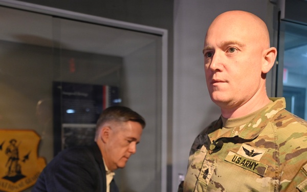 A U.S. Army soldier in OCP (Operational Camouflage Pattern) uniform stands in the foreground inside what appears to be a military heritage or display room. A second individual in civilian attire is visible in the background, leaning over a display table. Military memorabilia, including a leather flight jacket and a unit shield/crest, are visible in the background.