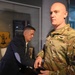 A U.S. Army soldier in OCP (Operational Camouflage Pattern) uniform stands in the foreground inside what appears to be a military heritage or display room. A second individual in civilian attire is visible in the background, leaning over a display table. Military memorabilia, including a leather flight jacket and a unit shield/crest, are visible in the background.