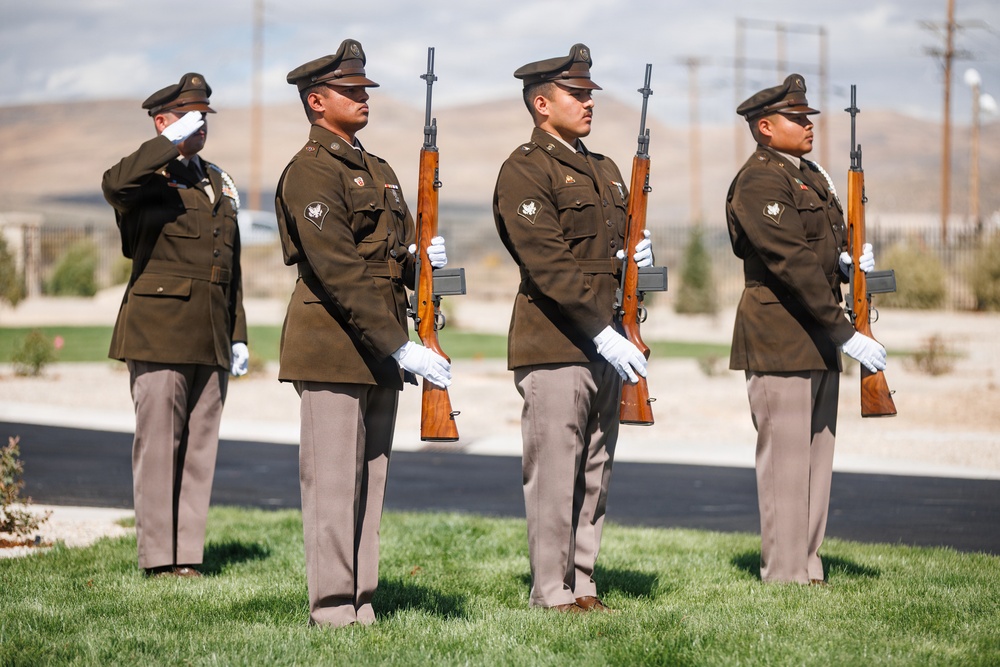 Four military personnel in dress uniforms are participating in a formal honor guard ceremony, likely at a cemetery or memorial site. Three soldiers stand at attention holding rifles while a fourth performs a salute in the background. The setting appears to be outdoors with a manicured lawn, arid landscape, and utility poles visible in the background.