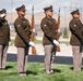 Four military personnel in dress uniforms are participating in a formal honor guard ceremony, likely at a cemetery or memorial site. Three soldiers stand at attention holding rifles while a fourth performs a salute in the background. The setting appears to be outdoors with a manicured lawn, arid landscape, and utility poles visible in the background.