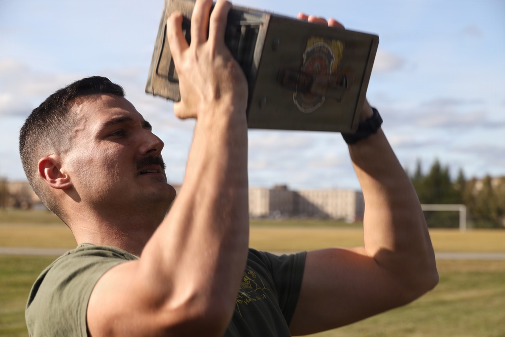 A man wearing a green military-style t-shirt is lifting a metal ammunition can (ammo can) overhead with both hands, appearing to perform a physical fitness exercise. He has a mustache, short hair, and is visibly sweating, suggesting intense physical exertion. The setting appears to be an outdoor athletic field or military installation.