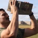 A man wearing a green military-style t-shirt is lifting a metal ammunition can (ammo can) overhead with both hands, appearing to perform a physical fitness exercise. He has a mustache, short hair, and is visibly sweating, suggesting intense physical exertion. The setting appears to be an outdoor athletic field or military installation.