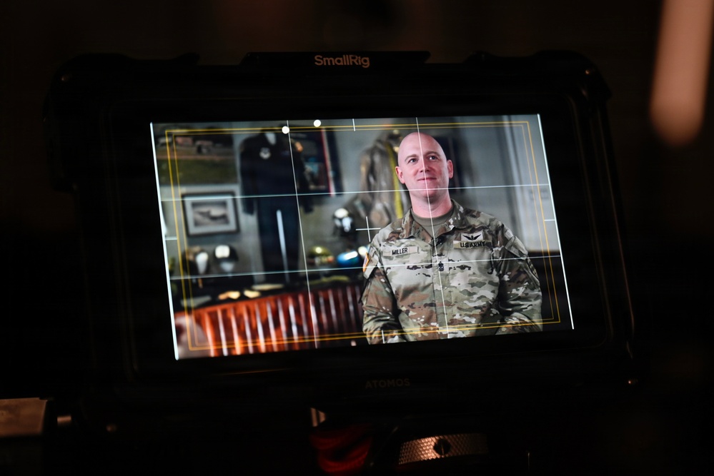A SmallRig/Atomos external monitor displays a U.S. Army soldier named Miller being filmed in what appears to be an official interview or video production setting. The soldier is wearing OCP (Operational Camouflage Pattern) uniform with a U.S. Army tape and a name tape reading 'MILLER', and rank insignia visible on the chest. The background on the monitor shows a formal office or display room with framed artwork, military memorabilia, and wooden furniture.