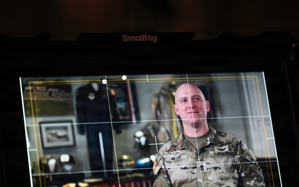 A SmallRig/Atomos external monitor displays a U.S. Army soldier named Miller being filmed in what appears to be an official interview or video production setting. The soldier is wearing OCP (Operational Camouflage Pattern) uniform with a U.S. Army tape and a name tape reading 'MILLER', and rank insignia visible on the chest. The background on the monitor shows a formal office or display room with framed artwork, military memorabilia, and wooden furniture.