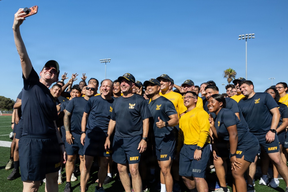 A large group of U.S. Navy personnel in uniform athletic wear (navy blue and gold shirts/shorts with 'NAVY' branding) are gathered on a sports field for a group selfie. One person on the far left is holding up a smartphone with a PopSocket to capture the photo. The group appears to be in high spirits, smiling and making gestures toward the camera.