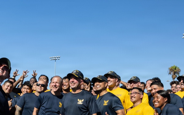 A large group of U.S. Navy personnel in uniform athletic wear (navy blue and gold shirts/shorts with 'NAVY' branding) are gathered on a sports field for a group selfie. One person on the far left is holding up a smartphone with a PopSocket to capture the photo. The group appears to be in high spirits, smiling and making gestures toward the camera.