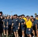 A large group of U.S. Navy personnel in uniform athletic wear (navy blue and gold shirts/shorts with 'NAVY' branding) are gathered on a sports field for a group selfie. One person on the far left is holding up a smartphone with a PopSocket to capture the photo. The group appears to be in high spirits, smiling and making gestures toward the camera.