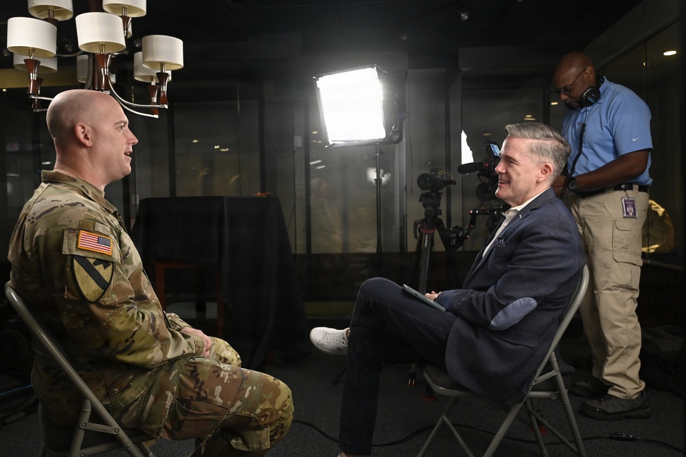 A U.S. Army soldier in camouflage uniform is being interviewed by a civilian in a navy blazer in what appears to be a professional media studio setting. A camera operator wearing headphones stands behind them, managing recording equipment. The setup includes professional lighting equipment and a camera on a tripod.