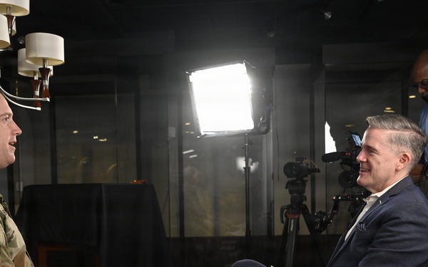 A U.S. Army soldier in camouflage uniform is being interviewed by a civilian in a navy blazer in what appears to be a professional media studio setting. A camera operator wearing headphones stands behind them, managing recording equipment. The setup includes professional lighting equipment and a camera on a tripod.