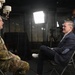 A U.S. Army soldier in camouflage uniform is being interviewed by a civilian in a navy blazer in what appears to be a professional media studio setting. A camera operator wearing headphones stands behind them, managing recording equipment. The setup includes professional lighting equipment and a camera on a tripod.