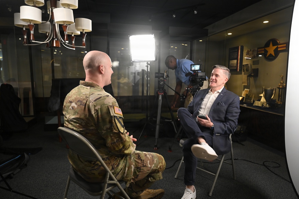 A military personnel in U.S. Army combat uniform is being interviewed by a civilian man in a navy suit, seated facing each other on folding chairs. A camera operator works behind them adjusting professional video equipment. The setting appears to be a military facility or museum, with display cases containing trophies and memorabilia visible in the background.