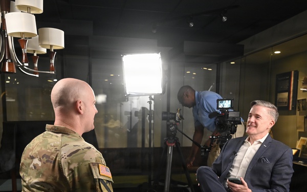 A military personnel in U.S. Army combat uniform is being interviewed by a civilian man in a navy suit, seated facing each other on folding chairs. A camera operator works behind them adjusting professional video equipment. The setting appears to be a military facility or museum, with display cases containing trophies and memorabilia visible in the background.