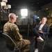 A military personnel in U.S. Army combat uniform is being interviewed by a civilian man in a navy suit, seated facing each other on folding chairs. A camera operator works behind them adjusting professional video equipment. The setting appears to be a military facility or museum, with display cases containing trophies and memorabilia visible in the background.