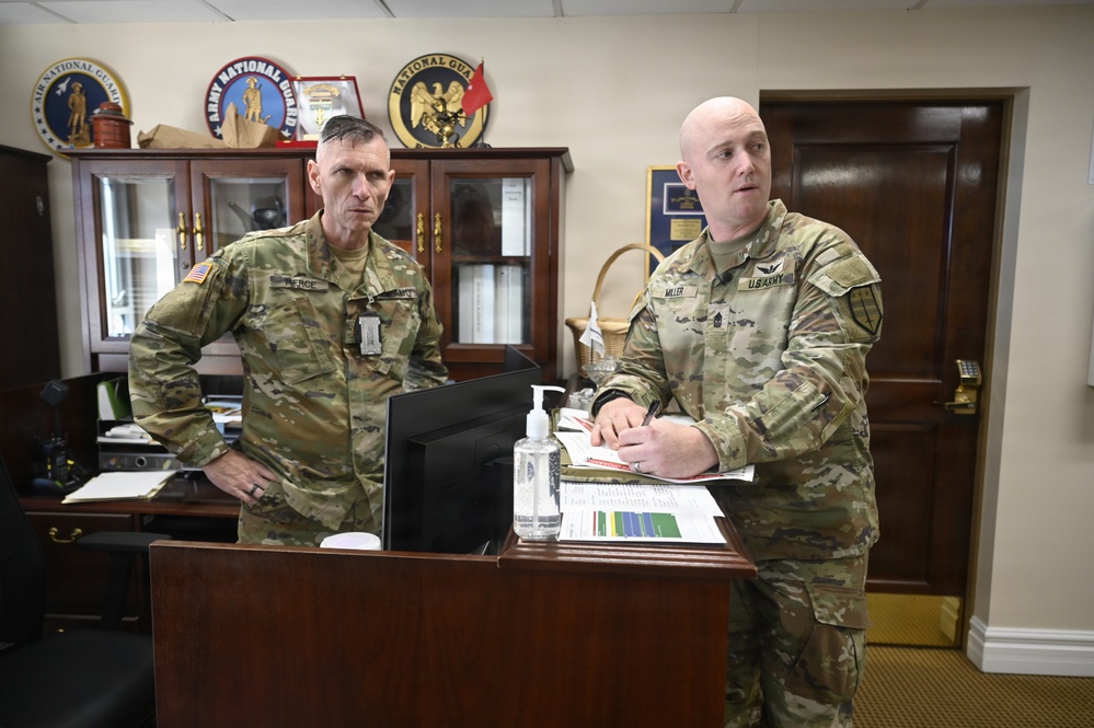 Two U.S. Army soldiers in OCP (Operational Camouflage Pattern) uniforms are standing at a desk in a military office environment. The soldier on the right, identified by his name tape as 'MILLER,' appears to be briefing or reviewing documents, while the soldier on the left, identified as 'PIERCE,' observes. The office background displays multiple military unit insignia plaques including Army National Guard and Air National Guard seals.