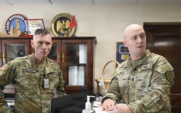 Two U.S. Army soldiers in OCP (Operational Camouflage Pattern) uniforms are standing at a desk in a military office environment. The soldier on the right, identified by his name tape as 'MILLER,' appears to be briefing or reviewing documents, while the soldier on the left, identified as 'PIERCE,' observes. The office background displays multiple military unit insignia plaques including Army National Guard and Air National Guard seals.
