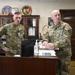 Two U.S. Army soldiers in OCP (Operational Camouflage Pattern) uniforms are standing at a desk in a military office environment. The soldier on the right, identified by his name tape as 'MILLER,' appears to be briefing or reviewing documents, while the soldier on the left, identified as 'PIERCE,' observes. The office background displays multiple military unit insignia plaques including Army National Guard and Air National Guard seals.