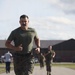 A male service member in U.S. Marine Corps MARPAT camouflage utilities and olive drab t-shirt runs at the front of a group on a marked track or road. Additional runners in similar military attire are visible in the background, suggesting a physical fitness test or training run. The setting appears to be a military installation with buildings and a parking area visible.